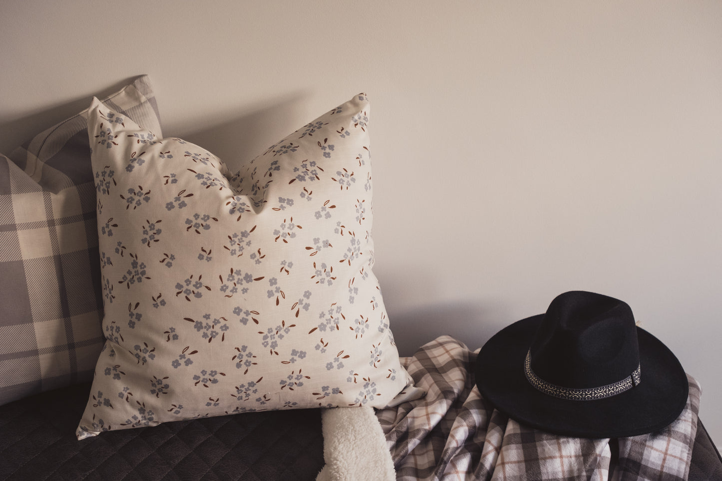 Image 4, the hand painted Wild petal Cushion by Lil & Dot in Dusky Blue on a bench with a soft throw blanket and a hat.