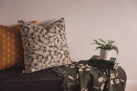 The Lil & Dot hand painted Daisy Cushion with the Soft Boom cushion on a bench with a woven blanket, books and a plant.
