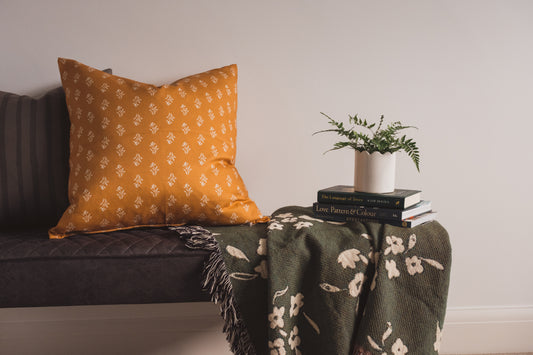 The hand painted Soft Bloom Cushion in Ochre and the Pinted strpe cushion by Lil & Dot on a bench with a woven green throw and a fern plant.