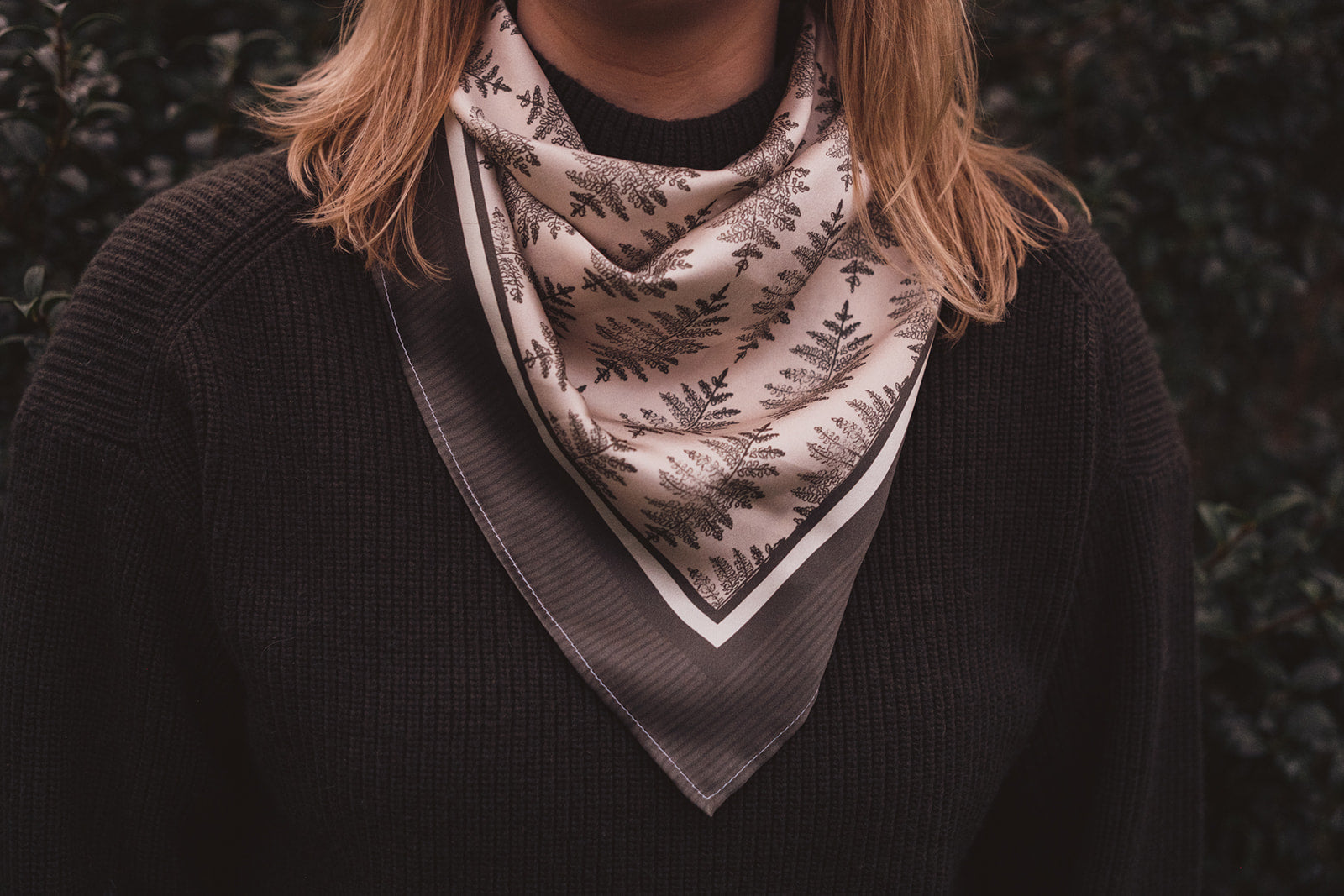 Image 2. A woman in a green knitted jumper wearing the hand illustrated Lil & Dot Fern Silk Bandana.
