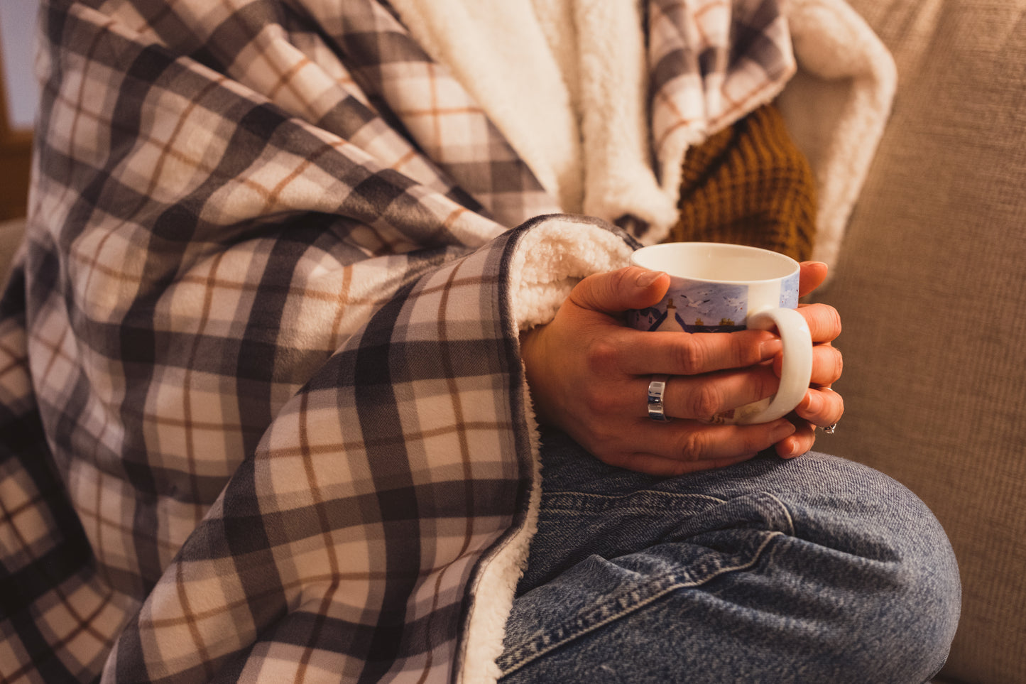 A woman with the Lil & Dot Soft Velvet Mocha Plaid Throw draped over her shoulders, with her feet up on the sofa with a cup of tea.