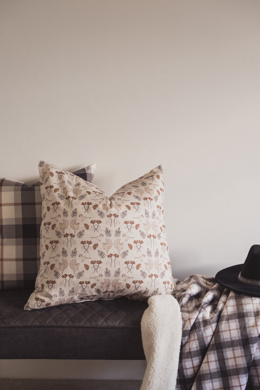 The Lil & Dot hand illustrated Birdsong Cushion in Mocha on a bench with a soft blue throw.